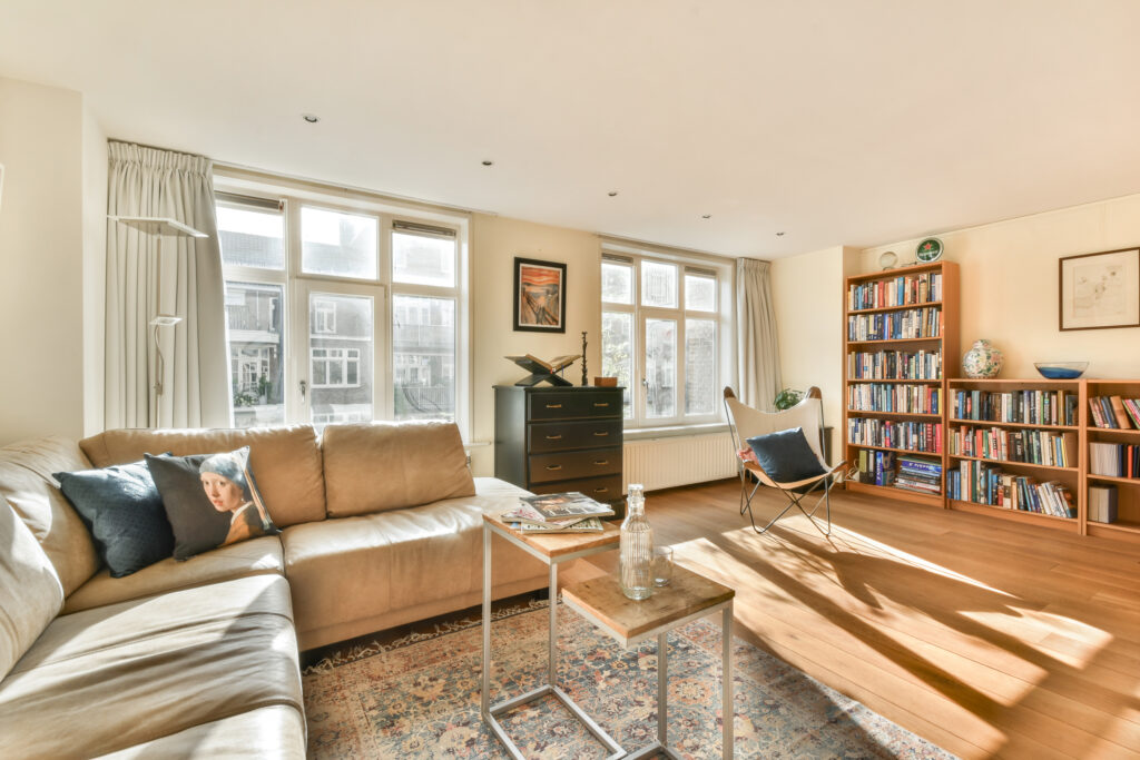 A spacious living room featuring large windows, a comfy sofa, and a rich variety of books on the shelves, creating a warm and inviting atmosphere.